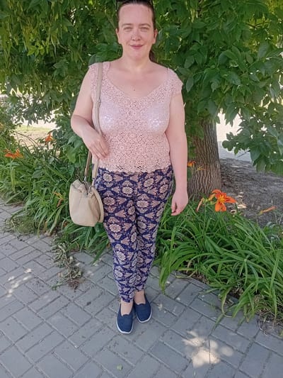 A woman stands outdoors on a paved walkway in front of a tree and garden. She is wearing a light pink, crocheted, short-sleeved top with a v-neck, and dark blue, patterned leggings. A light beige purse with a long strap is slung over her left shoulder. She has dark brown hair pulled back and is looking at the camera with a slight smile. Orange lilies are visible in the garden behind her, and the tree has green leaves. The lighting suggests it is daytime. This image was taken in Kherson, Ukraine.