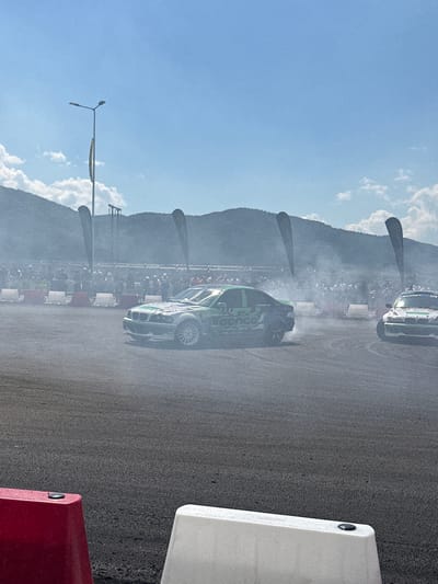 In Dospey, Bulgaria, on a sunny day with a clear blue sky and scattered clouds, a drift racing event is underway. Two race cars are visible on an asphalt track, engulfed in thick clouds of white smoke, indicating they are in the midst of drifting maneuvers. One car, predominantly green and white, with the text "Conce" prominently displayed on its side, is positioned towards the left of the frame. The second car, white with green and red accents and the number "176" on its side, is on the right. A crowd of spectators is lined up behind a barrier of white and red traffic cones and metal fencing, observing the race. In the background, a line of tall, dark banners stands against a backdrop of rolling, tree-covered hills. A tall lamppost with two lights is also visible to the left, behind the crowd. The foreground features two large, brightly colored traffic barriers, one red on the left and a white one on the right.