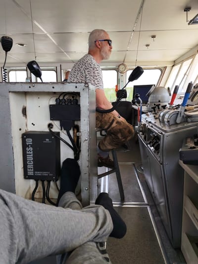 The image appears to be taken inside the wheelhouse of a vessel, possibly a boat or ship. One person, an older Caucasian male with a white beard and sunglasses, is seated on a stool and is turned to his right, looking out of the window. He is wearing a patterned shirt and brown pants. In the foreground, there are two legs clad in gray sweatpants, with black socks on the feet, that are extended toward the viewer, seemingly resting. There is an electrical box on the left of the image with the brand "UNICONT HERCULES-10" and the text "POWER SUPPLY 220AC/24DC 10A" on it. To the right of the seated male, there's a large control panel with various levers and switches. Microphones are hanging from the ceiling.

The location cannot be precisely determined as being in Khoroshevo, Russia based on the information in the image. There are no definitive visual elements in the photograph that specifically establish the city as Khoroshevo.