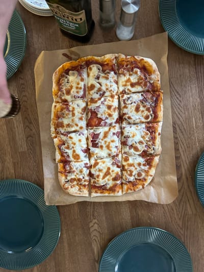 A rectangular pizza, cut into nine squares, lies on a piece of brown parchment paper on a wooden table. The pizza is topped with melted cheese, tomato sauce, and what appear to be mushrooms and sliced meat. Several dark green plates with ribbed edges are placed around the pizza. In the background, a bottle of extra virgin olive oil and salt and pepper shakers are visible. The overall scene suggests a meal setting, likely a casual gathering. There are no explicit indications of the location being Shepelyovo, Russia, as the image shows an indoor domestic setting with common dining items.
