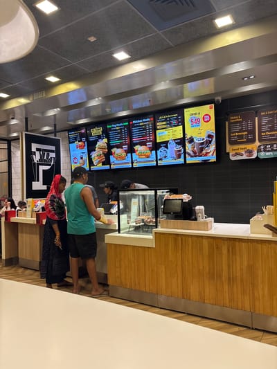 The image shows the interior of a McDonald's restaurant in Ambernath, India. Customers are standing in line at the counter, where an employee is taking an order. The menu is displayed on digital screens behind the counter, featuring various food and beverage options. The restaurant has a modern design with wood paneling on the counter and tiled walls. There is signage indicating the ordering area.
