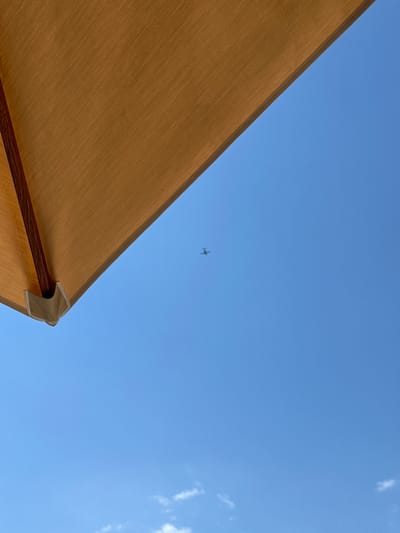 A low-angle view looks up at a clear, blue sky. In the upper left quadrant of the image, a section of a tan-colored umbrella is visible, casting a partial shadow. The umbrella appears to be made of fabric stretched over a wooden frame, with a visible wooden pole and white fabric corner reinforcement. In the center of the sky, a small, dark silhouette of an airplane is visible in flight. The bottom edge of the image shows a sliver of white clouds against the blue sky. The scene is set in Donja Lastva, Montenegro.