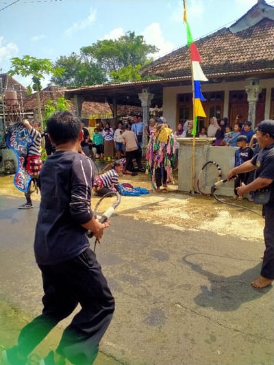 A group of people are gathered in front of a house in Kecamatan Kedawung, Indonesia (latitude: -7.52, longitude: 111.028).  A man dressed in a colorful costume, a black and blue costume, and a man wearing a striped shirt are performing in the street in front of the house. The man in the striped shirt is lying on the ground, while the other two men are standing. In the background, a group of people are watching the performance. A man in black is holding a whip. The house is made of brick and has a tiled roof. There is a flagpole with a colorful flag attached to it. The flag is a Buddhist flag.  There is a wooden gate to the left of the house. The street is paved with asphalt and has a dirt shoulder.  The sky is clear and the sun is shining.  There are trees and vegetation in the background.
