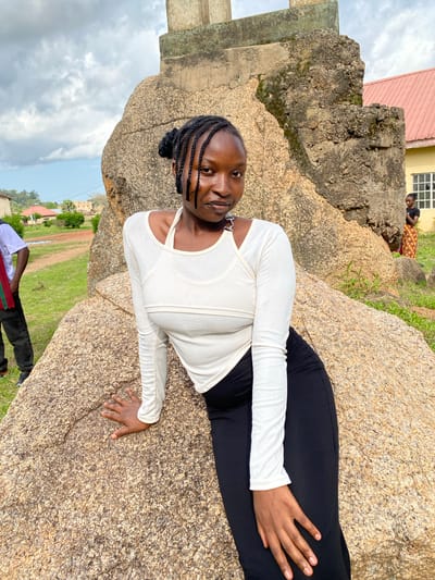A young Black woman with braided hair is leaning on a large boulder outdoors. She is wearing a white long-sleeved crop top and black pants. She is looking at the camera with a slight smile. In the background, there are buildings and trees. One building appears to be a school or administrative building with a red roof. Another person, wearing a white shirt and black pants, is standing to the left of the main subject. Further in the background, on the right, another young Black woman is visible near the building. The sky is cloudy. The environment appears to be a public space or park within a city. The scene is likely located in the city of Wang, Nigeria, given the context.