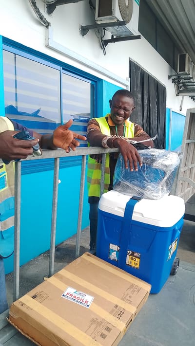 In Orerokpe, Nigeria, a man wearing a high-visibility vest laughs heartily as he leans on a metal railing. He is standing next to a blue and white Igloo cooler, which has a blue strap across its lid. On top of the cooler, there is a package wrapped in clear plastic. In the foreground, a brown cardboard box with "FRAGILE HANDLE WITH CARE" printed on a white sticker rests on the ground. The background features a building with bright blue siding and windows, and an air conditioning unit mounted on the wall. Another person's hands are visible on the railing to the left, with one hand gesturing outwards.