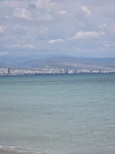 The image presents a wide coastal landscape. The foreground consists of a narrow strip of light-colored sand and shallow water with minor foam at the bottom edge, transitioning into a broad body of water exhibiting a greenish-blue hue and subtle surface ripples. In the midground, a city skyline extends horizontally across the horizon line. This urban area comprises numerous white and light-colored buildings, with several prominent modern high-rise structures visible, including a dark cylindrical tower on the left and multiple contemporary skyscrapers, some with curvilinear designs, toward the center-right. Behind the city, a range of hazy, bluish-grey mountains with sparse vegetation stretches across the background. The upper half of the image is occupied by a sky containing scattered, irregularly shaped grey and white clouds. No people, animals, or vehicles are present. The location depicted is the city of Unknown, Sovereign Base Areas of Akrotiri and Dhekelia.