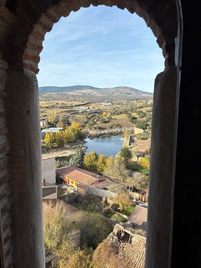 The scene presents an elevated panoramic view of Buitrago del Lozoya, Spain, framed through a weathered stone and reddish-brown brick archway. The interior edge of the arch is dark, contrasting with the brightly illuminated exterior. Beyond the arch, a calm body of water, consistent with a section of the Lozoya River or reservoir, dominates the midground, reflecting a clear blue sky. Its irregular banks are heavily vegetated with trees displaying vibrant yellow and orange autumn foliage, interspersed with darker green trees.
On the left side, below the arch, residential buildings with reddish-brown tiled roofs are visible. One prominent building has a light-colored facade with red accents. Further down and to the center-right, another building shows evidence of a significant roof collapse, with exposed wooden beams and scattered broken tiles. Along the water's edge on the right, several stone structures, possibly remnants of fortifications or older buildings, are present.
The background features undulating hills and distant mountains under a mostly clear blue sky with minimal scattered white clouds. The terrain between the town and the mountains appears open and somewhat arid, with sparse vegetation and a few indistinct scattered structures. No individuals are visible, and no specific actions are depicted. The overall environment suggests a sunny, clear autumn day.