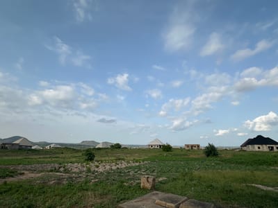 This is a daytime, eye-level, wide-angle photograph taken in Gidan Waziri, Nigeria, showcasing a rural landscape under a partly cloudy sky. The foreground features a rough, uneven terrain with patches of dry earth, rocks, and some low-lying green vegetation. Several large, square concrete blocks are visible in the immediate foreground, suggesting an unfinished construction or discarded building materials.

The midground is dominated by a sprawling, predominantly green field, with some areas appearing to be under cultivation with small, orderly rows of plants. Scattered throughout this area are several buildings in various stages of construction. To the left, a large, unfinished building with a peaked roof structure is prominent, and further back, several other houses with similar peaked roofs are visible, some appearing complete and others under construction. One building in the mid-right of the frame has a dark, steeply pitched roof.

In the background, rolling hills and mountains form the horizon. A hazy, distant cityscape or settlement can be observed between the hills. The sky above is a bright blue, punctuated by scattered white, fluffy cumulus clouds. The overall impression is one of development and expansion in a semi-arid rural setting. There are no people or animals visible in the image.