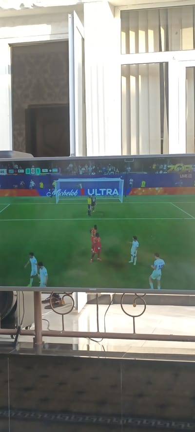 The image shows a television screen displaying a soccer match, likely viewed from a distance, suggesting an indoor setting in Baghdad, Uzbekistan. The screen exhibits a score graphic at the top left, indicating "COD 0 - 1 LTK". Below the score, a soccer field with players in red and white uniforms is visible, with a goal net and advertising boards featuring "Michelob ULTRA". The surrounding environment shows elements of an indoor space, possibly a living room or a cafe, with sunlight filtering through window blinds. The perspective suggests the viewer is seated or standing, with a railing or furniture in the foreground. The overall scene is characterized by diffused lighting and a slightly blurred quality, typical of an image captured of a screen.