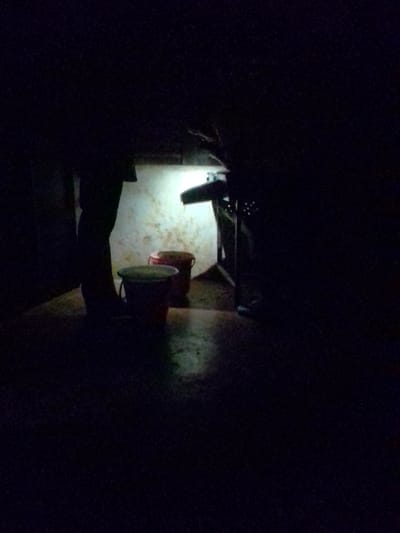 The image depicts a dimly lit scene, likely an outdoor area, illuminated by a bright light source from the upper center, casting strong shadows. In the foreground, a person's legs and feet are visible, standing on a textured surface that appears to be concrete or dirt. To the left of the person, two buckets are present. One is a dark-colored bucket with a light-colored lid, and it appears to be filled with a greenish substance. Behind this bucket, there is a red bucket. The background features a textured wall or surface, also illuminated by the light, and to the right, a dark, indistinct structure that could be a table or shelf, with some small dots of light suggesting it may have holes or be perforated. The overall environment suggests a utilitarian or storage area, possibly a workshop or an outdoor utility space in Borabu, Chitago Ward, Kenya.