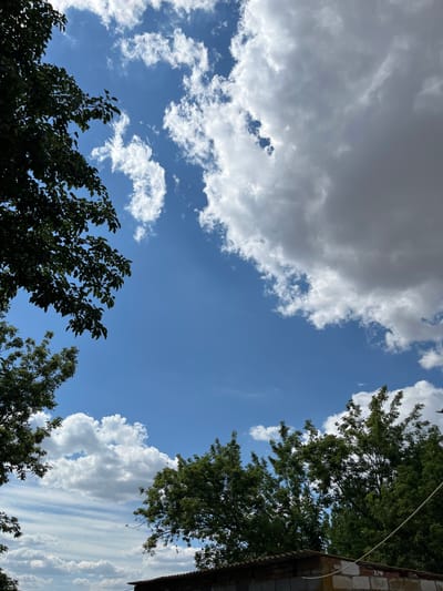 The image shows a view of the sky from the ground, with trees framing the edges of the frame. The sky is a clear blue with scattered white clouds, some of which are large and fluffy, while others are thin and wispy. The sun appears to be shining, casting light on the clouds. The trees are green and leafy, with branches reaching into the sky. In the foreground, a low structure made of bricks and corrugated roofing is visible. The setting appears to be outdoors in a rural or suburban area, possibly in Klynkyne, Ukraine, given the provided context. No people or animals are visible in the image. The overall atmosphere is bright and sunny, suggesting a pleasant day.