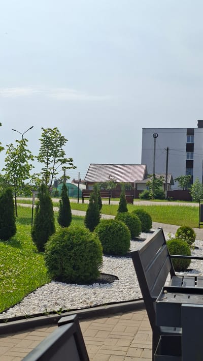 The image depicts an outdoor scene in the city of Smolevichi, Belarus. In the foreground, a modern dark gray bench with metal slats is positioned on a paved walkway made of light brown rectangular bricks. The bench faces towards a landscaped area featuring a border of white decorative stones. Behind the stones, a line of meticulously trimmed spherical green bushes is visible, interspersed with conical green evergreen trees. To the left of the bushes and trees, a patch of green grass dotted with small white flowers extends towards a paved path. Further into the background, a building with a white facade and multiple windows is situated. Adjacent to this building, a smaller structure with a corrugated metal roof, likely a house or shed, is visible. A street lamp with two lights stands to the left of the trees. The sky above is pale blue with sparse white clouds. The overall impression is of a well-maintained urban public space.