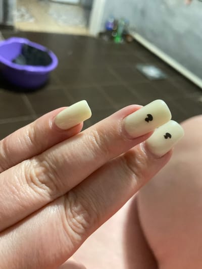 A close-up, high-angle shot shows a person's hand with artificial fingernails. The nails are a pale yellow color and are square-shaped with rounded edges. Two of the nails, on the middle and ring fingers, have small black heart shapes painted on them. The hand is resting on a surface that appears to be skin, likely a leg, due to the soft lighting and contours. In the background, out of focus, are a purple plastic basin and a dark, tiled floor. The overall environment suggests an indoor setting, possibly a home or a salon. The location is in the city of Rozkvit, Ukraine.
