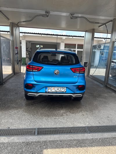 This media file features a rear view of a bright blue MG ZS car parked in an open-air car wash bay. The car is the central subject, positioned slightly to the right of the frame. Visible text on the car's license plate reads "PG LH275" and "JADRAN AUTO". To the left of the car, a metallic arm with a hose attached extends from the wall, suggesting it's part of the car washing equipment. In the background, through glass panels, we can see parts of the car wash facility, including signs with text such as "TEPIH SERVIS TIP TOP" and what appears to be an air conditioning unit. The ground is a concrete surface with a drainage grate running along the right side of the image. The lighting suggests it is daytime, likely sunny, casting shadows. The scene depicts a common urban or suburban setting, specifically a self-service car wash. There are no people visible in the image. The overall impression is one of a car being prepared for or recently having been washed.