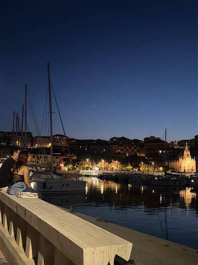 This image captures a nighttime scene in the city of Imperia, Italy, likely at the Imperia Oneglia harbor. In the foreground, a couple is seated on a stone ledge, facing away from the camera and towards the water and the cityscape. The woman has long, dark hair and is wearing a black top and blue jeans. The man has dark hair and is wearing a black shirt. They appear to be in conversation or enjoying the view together.
The background features a harbor filled with docked sailboats and smaller vessels. The masts of the sailboats create a strong vertical element against the skyline. Beyond the harbor, the city of Imperia unfolds, with buildings illuminated by warm lights, creating a picturesque backdrop. A prominent church with a tall spire stands out among the buildings, its facade brightly lit. The dark blue sky above is clear, with a few faint stars visible. The calm water of the harbor reflects the lights of the city and the boats, adding a shimmering quality to the scene. The overall atmosphere is peaceful and romantic, indicative of an evening stroll along the Italian coast.