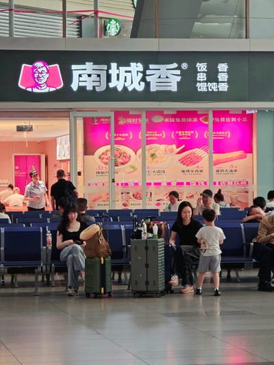 The image captures a scene inside a restaurant in Fengtai District, China. A large sign above the restaurant entrance displays "南城香" (Nan Cheng Xiang) in white lettering, accompanied by a pink cartoonish portrait of a man with glasses. Below the main sign, a promotional banner features images of various food items, including dumplings, skewers, and bread, with prices listed in Chinese Yuan.
Several individuals are seated on blue benches in what appears to be a waiting area or dining section. In the foreground, a woman in a black top and patterned pants sits with her legs crossed, holding a mobile phone and looking down. Next to her, a young boy in a white t-shirt and grey shorts stands facing away from the camera. A green suitcase is positioned beside the woman.
Further back, other people are seated, some interacting with each other, while others are looking in different directions. A woman wearing a white shirt and a cap, possibly an employee, is standing and gesturing with her hand near an open doorway. The environment is well-lit, with reflections visible on the tiled floor. The overall impression is one of a busy public eatery, likely a food court or a fast-food restaurant within a larger establishment like a train station or shopping mall.