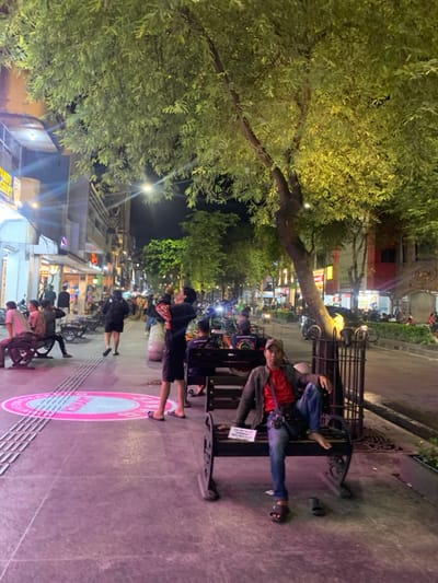 This image depicts Malioboro Street in Yogyakarta, Indonesia, at night. The scene is illuminated by streetlights and shop lights, casting a mix of bright and shadowed areas.
In the foreground, a wide, paved pedestrian sidewalk extends from left to right, featuring tactile paving strips. A circular pink graphic is visible on the sidewalk, with text partially readable as "SELAMAT DATANG" (Welcome).
On the right side of the foreground, a man sits on an ornate dark metal bench. He wears a dark jacket over a red t-shirt, blue jeans, and a light-colored baseball cap. His left foot is in a dark sandal, while his right foot is bare and rests on the bench. A dark shoulder bag is visible across his body, and he holds a small white rectangular sign. Directly behind him, a tree with a gnarled trunk and lush green foliage stands, illuminated by an upward light source.
Towards the mid-ground, slightly to the left of the center, another man stands facing right, looking upwards, with his arms raised as if holding a device to photograph or film something above him. He wears a dark t-shirt and dark shorts, with light-colored flip-flops. Further left, several other individuals are present, some sitting on benches or parts of what appear to be traditional becak (cycle-rickshaws).
The background features a row of multi-story commercial buildings lining the street, displaying illuminated signs such as "PANDU SHIRT." A multi-lane road runs parallel to the sidewalk, populated by vehicles, including cars and motorcycles, and additional pedestrians. More illuminated trees are visible further down the street. Overhead, streetlights emit bright light into the dark sky.