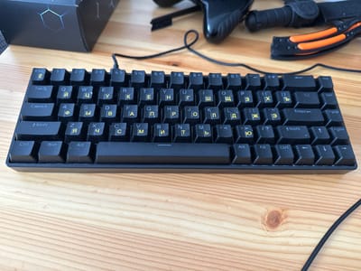 The image displays a black mechanical keyboard with yellow Cyrillic lettering on a light-colored wooden desk. The keyboard is positioned horizontally, with the top edge closer to the viewer. Keycaps are visible, showing letters such as 'E', 'Y', 'U', 'I', 'O', 'P', 'A', 'S', 'D', 'F', 'G', 'H', 'J', 'K', 'L', 'Z', 'X', 'C', 'V', 'B', 'N', 'M', and numbers and symbols along the top row. Modifier keys like 'Shift', 'Ctrl', 'Win', and 'Alt' are also present. A black USB cable extends from the top left of the keyboard and disappears off-camera. In the background, partially visible objects suggest a gaming setup: a black box with a hexagonal pattern on its lid and a black gaming controller with orange accents. The lighting suggests an indoor environment, likely a home office or gaming area. There is no discernible information to place this image in Zielona Góra, Poland.