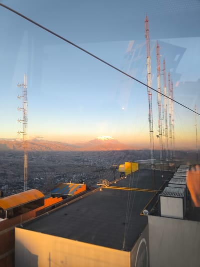 The image is an aerial view of El Alto, Bolivia, at sunset, taken from a cable car. In the foreground, there are several buildings with flat roofs, including one with an orange, curved roof structure and another with a yellow awning. Various radio and television towers are prominent in the scene, with some in the foreground and others stretching into the distance. The city of El Alto sprawls across the landscape, appearing as a dense collection of buildings. Beyond the city, rolling hills and mountains are visible, with the highest peak, likely Mount Illimani, covered in snow and illuminated by the setting sun. The sky is a gradient of blue and orange, indicating the time of day. A thin cable, likely part of the cable car system, runs diagonally across the upper portion of the image. There is a faint reflection on the glass, suggesting the photo was taken through a window. No people are visible in the image.