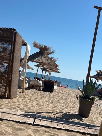 The image depicts a sunny day at a beach resort in Dyuni, Bulgaria. The foreground shows a wooden walkway laid on the sand, leading towards the sea. To the left, a woven structure with a thatched roof provides shade, with white drapes hanging down. Further inland, several straw-thatched umbrellas offer shelter, and large white beanbag chairs are scattered on the sand. In the background, the calm blue sea stretches to the horizon under a clear blue sky. A tall wooden pole stands to the right of the frame, and a large potted agave plant is placed near the edge of the walkway. Several people are visible, some relaxing on the beanbag chairs, others near the water, and at least one person is swimming. A red inflatable raft is also present in the sea. The overall scene conveys a relaxed and leisurely atmosphere typical of a beach vacation.