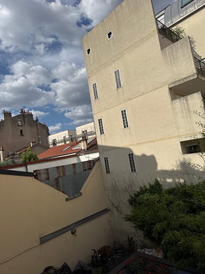 The image depicts a view of the urban environment in Montreuil, France, specifically looking up at buildings and rooftops from a slightly elevated perspective. In the foreground, a beige building with a textured facade dominates the right side of the frame. It features several rectangular windows, some with glass blocks and others with dark frames. Balconies with black metal railings are visible on the upper floors. To the left, a lower beige structure angles away, with a corrugated metal roof section visible at its peak.
Behind these structures, older buildings are visible. One prominent building on the left has a grey facade with multiple chimneys and windows. Further in the background, modern apartment buildings under construction, adorned with scaffolding, can be seen. Red-tiled roofs are interspersed among the buildings, adding a common architectural element. The sky above is partly cloudy, with dramatic white and grey clouds set against a blue sky. Lush green foliage and trees are present in the lower right quadrant of the image, partially obscuring the view of the ground level. On the ground, a bicycle and some covered items are visible, suggesting a residential or mixed-use area. The overall scene conveys a sense of a dense urban landscape with a mix of old and new construction, bathed in natural light under a dynamic sky.