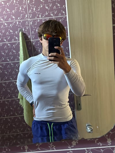 In a bathroom with purple floral wallpaper, a young man stands in front of a mirror, taking a selfie. He is wearing a white long-sleeved shirt with the brand name "NAIAJO" visible, and blue swim shorts with neon green drawstrings. He has on red and orange mirrored sunglasses, and his brown hair is messy. A light green towel hangs on a hook to his left. The mirror reflects the bathroom's tiled walls and a wooden door. The image's context places the scene in Vranjina, Montenegro.