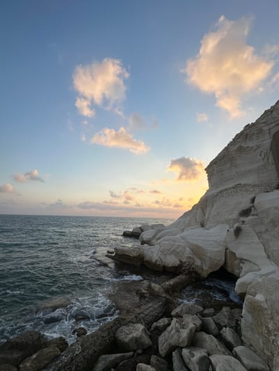 This image depicts a coastal scene at sunset in the city of Mate Asher Regional Council, Israel. The foreground features a rocky shoreline with waves gently crashing against the rocks. The water is a deep blue-green. To the right, a large, white cliff face rises dramatically, showing layered rock formations. The sky above is a soft blue with scattered clouds illuminated by the setting sun, casting a warm orange glow. The horizon line is visible where the sea meets the sky. The scene is devoid of people or any visible structures. The overall impression is one of natural beauty and tranquility.