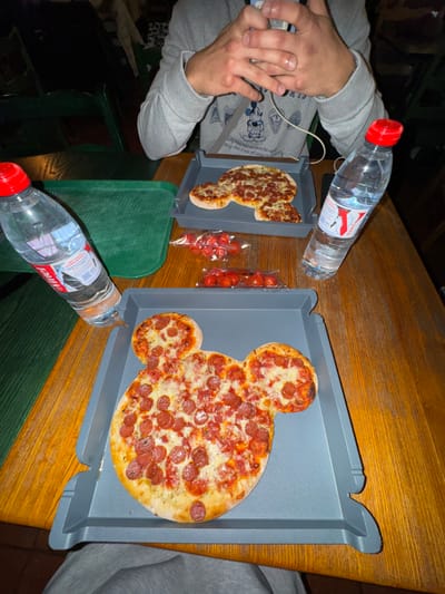 A wooden table displays two Mickey Mouse-shaped pizzas on gray trays. One pizza is whole, covered in pepperoni, and the other is partially eaten. Next to the smaller pizza are two small bags of cherry tomatoes. Two bottles of water with red caps are also on the table. A person wearing a gray sweatshirt with Mickey Mouse print is visible on the left, and another person wearing a gray sweater is on the right. Green wooden chairs surround the table.