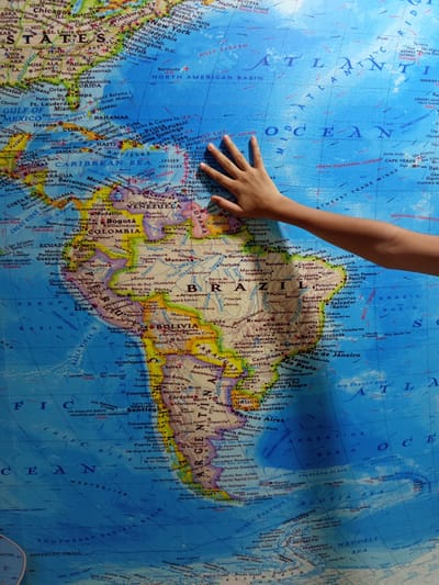 A person's hand is resting on a large map of the world. The map displays continents, oceans, and country borders. The hand is positioned over South America, specifically between Colombia and Venezuela, in the region of the Caribbean Sea. The city of Coveñas, Colombia, is not visible in this detail on the provided map. The background is neutral and does not provide any additional context.