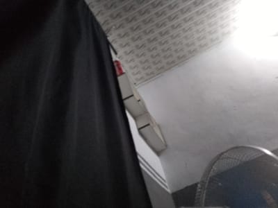 The image shows a close-up view of a black curtain or fabric on the left side, partially obscuring the background. The background consists of a white ceiling with a textured, patterned surface, possibly acoustic tiles, running horizontally across the top of the frame. Below the ceiling, a white wall is visible. In the lower right corner, the edge of a black fan with a metal grille is present. A sliver of a red object is visible behind the black fabric, near the ceiling. The lighting suggests an indoor environment with a light source illuminating the upper right portion of the scene. The image quality is grainy. This scene is not in Senti, Nigeria.