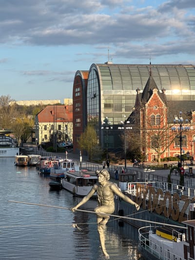 A bronze statue of a nude male figure poised as if diving into the water is the focal point of this image. The statue is located on the Brda River in Bydgoszcz, Poland. The riverbank is lined with docked boats, and a modern, curved building with a glass facade stands behind the statue, reflecting the sky. Adjacent to this modern structure is a traditional red brick building. People are seen walking along the riverbank. The sky is partially cloudy.