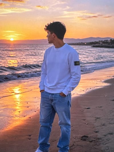 A young man stands on a sandy beach during sunset. He is wearing a white long-sleeved shirt with a dark logo on the left sleeve and light blue jeans. His hands are in his pockets, and he is looking out at the ocean. The sun is setting on the horizon, casting a warm orange glow over the water and the sky. Waves are gently rolling onto the shore. The beach is wet from the receding tide, and there are small pebbles and sand visible. In the distance, there are hills and some buildings. The scene is tranquil and serene. This image is not located in Neubrandenburg, Germany.