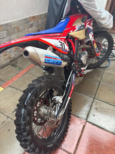 This media file shows a dirt bike parked on a wet tiled patio. The bike is red, white, and blue with black accents. The front wheel is partially covered by a white tarp. The rear wheel is exposed and shows a knobby tire with "MITAS" and "EXCEL" visible on the sidewall. The bike has a silver exhaust pipe with "D.I.D." written on it in blue letters. Red and white decals are visible on the bike's body. The ground is tiled with reddish-brown and grey tiles, and there are water puddles on the surface, indicating it has been raining or the area has been washed. The scene appears to be outdoors, possibly in a backyard or driveway. There are no people visible in the image. The lighting suggests it is daytime. The overall impression is that the bike is either recently cleaned or has been exposed to the elements.