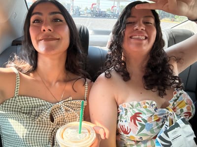 Two women are inside a car and appear to be taking a selfie while upside down. The woman on the left has curly brown hair and is wearing a floral patterned top. She is holding up a peace sign. The woman on the right has straight brown hair and is wearing a green and white checkered top. She is holding a clear plastic cup with a green straw. Outside the car window, various signs and vehicles are visible, but the exact location within Santa Ana Xalmimilulco, Mexico, cannot be determined from the image.