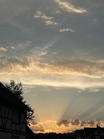 This is a photograph of a sunset over the town of Heinebach, Germany. The sky is filled with dramatic clouds, with rays of sunlight emanating from the horizon. In the foreground, a traditional half-timbered house is silhouetted against the vibrant sky. The house has dark beams and lighter infill, with shutters on the windows. To the left of the house, a tree with dark foliage is visible. The horizon is lined with trees, and in the far distance, another building with a dark roof is partially visible. The overall impression is one of a serene and picturesque evening.