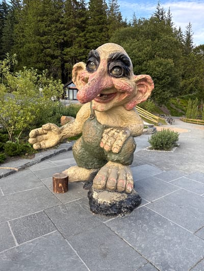 A life-sized, rough-textured statue of a troll, depicted in a seated position, is the central subject of the image. The troll has large, round, light-colored eyes with dark pupils and heavy black eyebrows. Its nose is long and bulbous, with prominent nostrils, and its mouth is open in a grin, revealing several teeth and a pink interior. Large, floppy ears frame its head, and its skin is a pale yellowish-brown, with reddish-pink accents on its nose, ears, and fingers. The troll is wearing green overalls, and its large hands are prominent, with wrinkled fingers and painted pink fingernails. One hand is extended outwards, palm up, while the other rests on its knee. It is positioned on a paved area of large, grey, rectangular flagstones, with a small wooden log serving as a makeshift stool or prop nearby.
The environment surrounding the troll suggests an outdoor park or attraction. Behind the statue, a dense forest of tall, green coniferous trees forms a natural backdrop. In the mid-ground, there is a small building with glass windows, possibly a visitor center or shop, and glimpses of playground equipment including wooden railings and slides. Further in the background, the terrain slopes upwards, covered with vegetation and trees, under a sky with scattered white clouds. The overall scene appears to be a tourist attraction, likely in a natural setting, though a specific location in Bergen, Norway, cannot be definitively determined from the image alone. The lighting suggests it is daytime, likely late afternoon or early evening, given the soft shadows.
