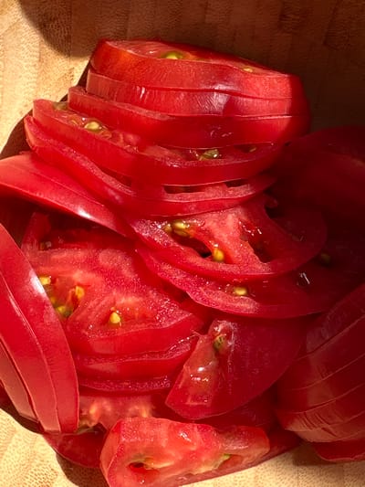 A close-up, overhead shot shows sliced tomatoes in a wooden bowl. The tomatoes are bright red and appear fresh, with visible seeds and pulp. Some slices are stacked neatly, while others are more haphazardly arranged, creating a layered effect. The wooden bowl has a textured grain and a warm, natural color. Sunlight casts a shadow on the left side of the bowl, creating a contrast between light and dark areas. There are no people or animals visible in the image, and no specific location in Novoaleksandrovo, Russia can be identified from the visual information. The scene is static and focused solely on the prepared tomatoes.