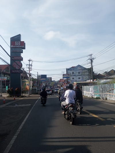 The image shows a street scene in what appears to be a busy area. A group of people are riding motorcycles down the road, facing away from the viewer. The road has a painted yellow line. On the left side of the road, there is a tall sign with multiple company logos, including "ciplaz Cipanas", "Ramayana", "Robinson mart", "GOKANA RAMEN & TEPPAN", and "MAKO". Further down the road, numerous signs and advertisements are visible, including a KFC sign. Buildings line both sides of the street, with power lines overhead. The sky is partly cloudy. The license plate on one of the motorcycles reads "B 3554 UXT 12-26." The exact location in Phou Khoun, Laos, cannot be determined from the image alone.