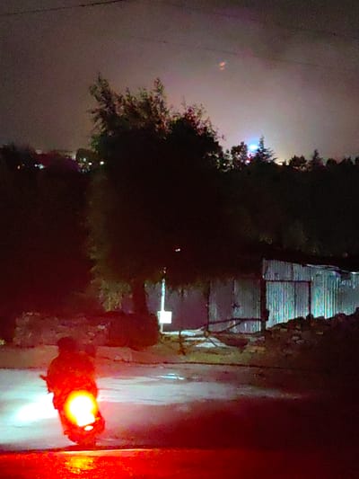 The image depicts a night scene in the city of Kulang, India, likely in a rural or semi-rural area. In the foreground, a person is silhouetted on a scooter, illuminated by the bright headlight and taillight, casting a strong red glow on the asphalt road. The road surface appears wet, reflecting the lights.
Behind the scooter, several trees are visible, their foliage dark against the dimly lit sky. Beyond the trees, a corrugated metal structure, possibly a shed or small building, stands to the right. The structure has vertical lines from the corrugated metal and is partly obscured by shadows. In the far distance, on a hilltop or elevated area, a cluster of lights suggests a settlement or town. The sky is overcast and dark, with some faint streaks of color visible, possibly from distant artificial lights or atmospheric phenomena. A thin wire is stretched horizontally across the upper portion of the image. The overall atmosphere is dim and somewhat eerie, characteristic of a nighttime rural environment.