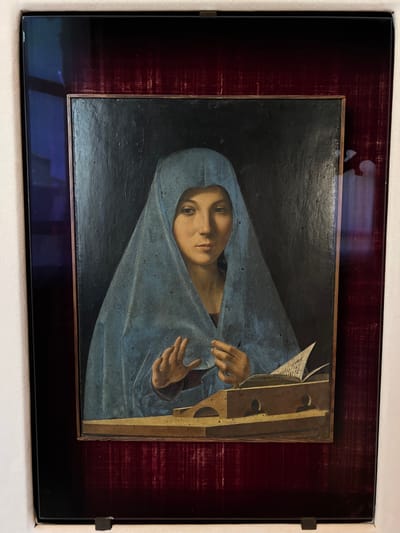 This is a photograph of a painting, likely displayed in a museum or gallery in Palermo, Italy. The painting depicts a close-up portrait of a person, possibly a woman, wearing a blue garment that drapes over her head and shoulders, obscuring much of her body. Her face is centrally positioned and illuminated, with a somber or contemplative expression. Her eyes are looking directly at the viewer. One hand is visible, with fingers delicately posed, appearing to interact with the fabric of her garment or an unseen object. To the right of the figure, a portion of a wooden object, perhaps a lectern or a piece of furniture, is visible. It has a textured surface and several circular indentations. A triangular piece of paper, possibly a sheet of music or a document, is propped against this object, with some faint markings that could be text. The background of the painting is dark and indistinct, creating a focus on the figure. The painting is framed and mounted within a larger, dark, reflective border, possibly a glass display case. The overall mood conveyed by the painting is one of quiet introspection and possibly religious devotion, given the common iconography of figures in blue veils in religious art. There is no indication of the time of day, weather, or specific location within Palermo beyond the context of its display.