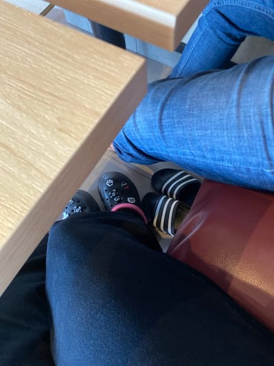 The image shows a low-angle view of people's legs and feet in what appears to be an indoor setting, possibly a cafe or restaurant, given the wooden table in the foreground and the red upholstered seating in the background. Two individuals are visible. One person is wearing blue jeans and black slides with white stripes. The other person is wearing dark pants and black Crocs adorned with various charms, including what look like paw prints and possibly other decorative elements. The lighting suggests it could be daytime. The scene is casual and relaxed, with people seemingly seated or about to be seated at a table. There is no visible text in the image. The location context provided is Lhoksukon, Indonesia, but there are no specific visual cues within the image to confirm this location.