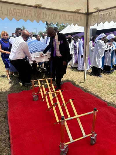 The scene takes place outdoors, at a funeral service in Pridelands, Kenya. The image shows a group of people attending a funeral service, standing under a tent in a grassy area. The image is focused on a coffin being carried by two men. The coffin is covered in a blue and white checkered cloth. The coffin is being carried towards a golden, foldable stretcher with wheels. The two men are wearing white shirts. One man is wearing a suit jacket. One man is wearing a dark suit with a purple tie. The other men in the background are standing behind the coffin and are dressed in various clothes. Some are wearing suits and others are wearing casual clothes. All of the people in the photo are facing the camera. There is a red carpet leading to the stretcher.  The tent reads “L HOME LTD MONITEZA AMUZAIA FUNEREAL” in reverse, which appears to be the company name in Swahili.