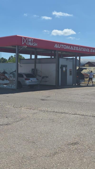 A person in a white shirt and dark shorts is seen standing next to a car at an automated car wash. The car wash has a large red awning with white text that reads "MY WASH" and "AUTOMAŠGĀTAVA 24". The scene is outdoors, under a bright blue sky with scattered white clouds, suggesting it is daytime and the weather is pleasant. The ground is paved with asphalt. The car appears to be a silver sedan with a license plate visible, though the text is not clearly legible. The car wash structure itself is modern, with metal supports and panels. There are also some colorful graphics on the wall behind the car, depicting water and car wash symbols. The overall impression is of a typical day at a self-service car wash.