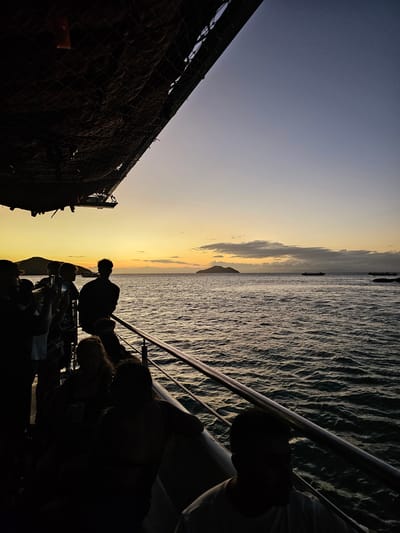 The image depicts a sunset scene from a boat in Armação Dos Búzios, Brazil. A group of silhouetted people is visible on the right side of the frame, standing near a railing. To the left of the people is a view of the ocean's surface, which reflects the colors of the sunset. In the distance, there are clouds and the silhouette of a mountain, possibly a small island, against the fading light. The sky transitions from a pale blue to shades of orange and yellow near the horizon. A structure covered with a net, possibly part of the boat, occupies the left side of the image.