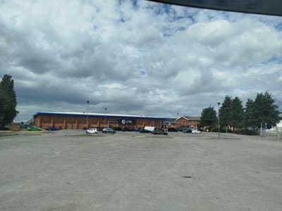 The image shows a wide shot of a large, empty car park in front of a JD Gyms building. The building is made of brick and has a blue roof. The car park is paved and appears to be quite large, with several cars parked in it. The sky is cloudy and overcast. The location is JD Gyms, 229 East Prescott Road, Liverpool, United Kingdom.