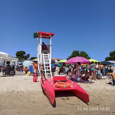 The image depicts a beach scene on a sunny day in Trani, Italy. A red lifeguard station with a white ladder stands prominently in the foreground, with the word "SALVATAGGIO" visible on its red canopy. A lifeguard wearing a red vest is seated at the top of the station. A red rescue boat with an orange life ring is positioned on the sand in front of the station. Beachgoers are visible in the background, some relaxing under umbrellas, others wading in the water. The environment is sandy, with scattered beach umbrellas and chairs. A person in a wheelchair is also present near the lifeguard station. The sky is clear and blue. A timestamp "22.06.2026 10:57" is overlaid on the bottom right of the image.