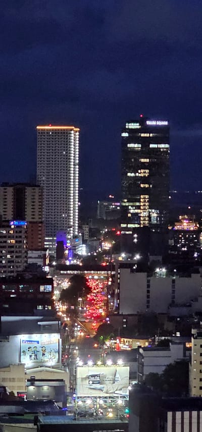 This is a nighttime aerial view of a city street in Makati, Philippines. The image is oriented vertically, showing a densely built urban landscape illuminated by artificial lights.

The primary subjects are buildings of various heights, including a prominent tall skyscraper with "TRIUM SQUARE" visible in bright lettering on its facade. Another large, multi-story building with rows of illuminated windows runs horizontally across the lower portion of the frame, its facade outlined with bright orange lights. Numerous other buildings, some appearing to be commercial or residential, are visible, characterized by their illuminated windows and rooftop lights.

The scene depicts a busy street at night, with a notable concentration of red and white lights suggesting heavy traffic, possibly a traffic jam or a vibrant street event. Streetlights and the lights from shops and vehicles contribute to the bustling atmosphere. Billboards are visible on some buildings, including one with text that appears to say "70 years of experience." Another billboard displays cartoonish figures and text, part of which reads "EVERY STEP OF THE WAY."

The time of day is clearly night, as indicated by the dark sky and the reliance on artificial illumination. The weather appears to be clear, as the lights are sharp and there are no visible signs of rain or fog. The location is identified as Makati, Philippines. The overall mood conveyed is one of urban activity and vibrant nightlife, with the glowing lights creating a lively and somewhat chaotic impression.