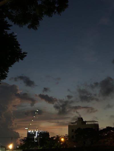 A twilight scene in Da Nang, Vietnam, captures a dusky sky with scattered clouds tinged with orange and pink hues from the setting sun. In the foreground, dark silhouettes of trees frame the upper left corner, with a single bright star or planet visible in the deep blue sky.
Mid-ground features a construction site with a tall crane illuminated by a string of lights that ascend its boom, over a dark, multi-story building under construction, also lit from within. To the right, a lighter-colored, low-rise building with a rooftop structure stands against the darkening sky, partially obscured by trees. The lower portion of the image shows more dark foliage and streetlights casting a warm glow. The overall impression is one of a city transitioning from day to night.