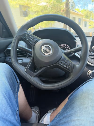 The image presents a first-person perspective from inside a modern Nissan vehicle, focusing on the steering wheel, parts of the dashboard, and the driver's lower body.
The central element is a black, multi-function Nissan steering wheel, featuring a silver Nissan logo at its core and "AIRBAG" text directly below it. On the left side of the steering wheel, a set of cruise control buttons is visible, including "RES+", "CANCEL," and "SET-." The right side houses controls for audio (volume up/down, track skip) and phone functions (answer/end calls). A wiper stalk, partially seen behind the steering wheel, shows an "AUTO" setting. Through the steering wheel, a portion of the instrument cluster, displaying white numbers on a dark background, is visible, along with an air vent.
The driver's legs are seen in the lower right portion of the frame. The driver wears light blue denim jeans, white ribbed socks, and light-colored sneakers. Patches of bare skin are visible on the shins between the jeans and socks.
Through the car's windshield, a bright outdoor environment is reflected. Strong sunlight illuminates a light-colored, multi-story building with several windows, partially framed by green trees and branches. This suggests a clear, sunny daytime setting in an urban or suburban area within Lawrence Township, United States of America.