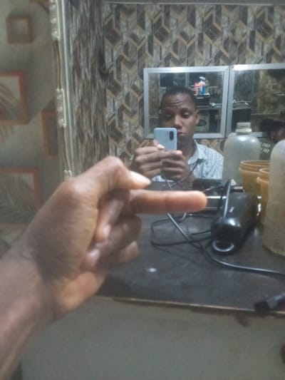 In Ijegun, Nigeria, a person is captured in a selfie in what appears to be a barber shop. The individual, holding a light blue smartphone, is positioned in front of a mirror, reflecting a light-colored checkered shirt. The background reveals multiple mirrors, toiletries including toothbrushes and toothpaste, a large plastic water dispenser, and stacks of yellow containers. A black electrical appliance, possibly a clipper, with wires is also visible on the dark countertop. In the foreground, the person's left hand is prominently displayed, with the middle finger extended towards the camera. The overall scene is dimly lit and suggests a casual, personal moment.